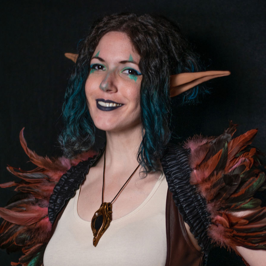 Nikki Crenshaw dressed as an elf. Her hair is black with a touch of green, and falls to the sides of her face in two long curls., with Elf ears sticking out of it. Her eyes covered by green sigils make-up. A wooden neclace hangs from her neck as she smiles.